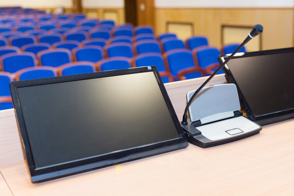 Conference podium with a mic and two display screens in front of rows of blue empty seats.